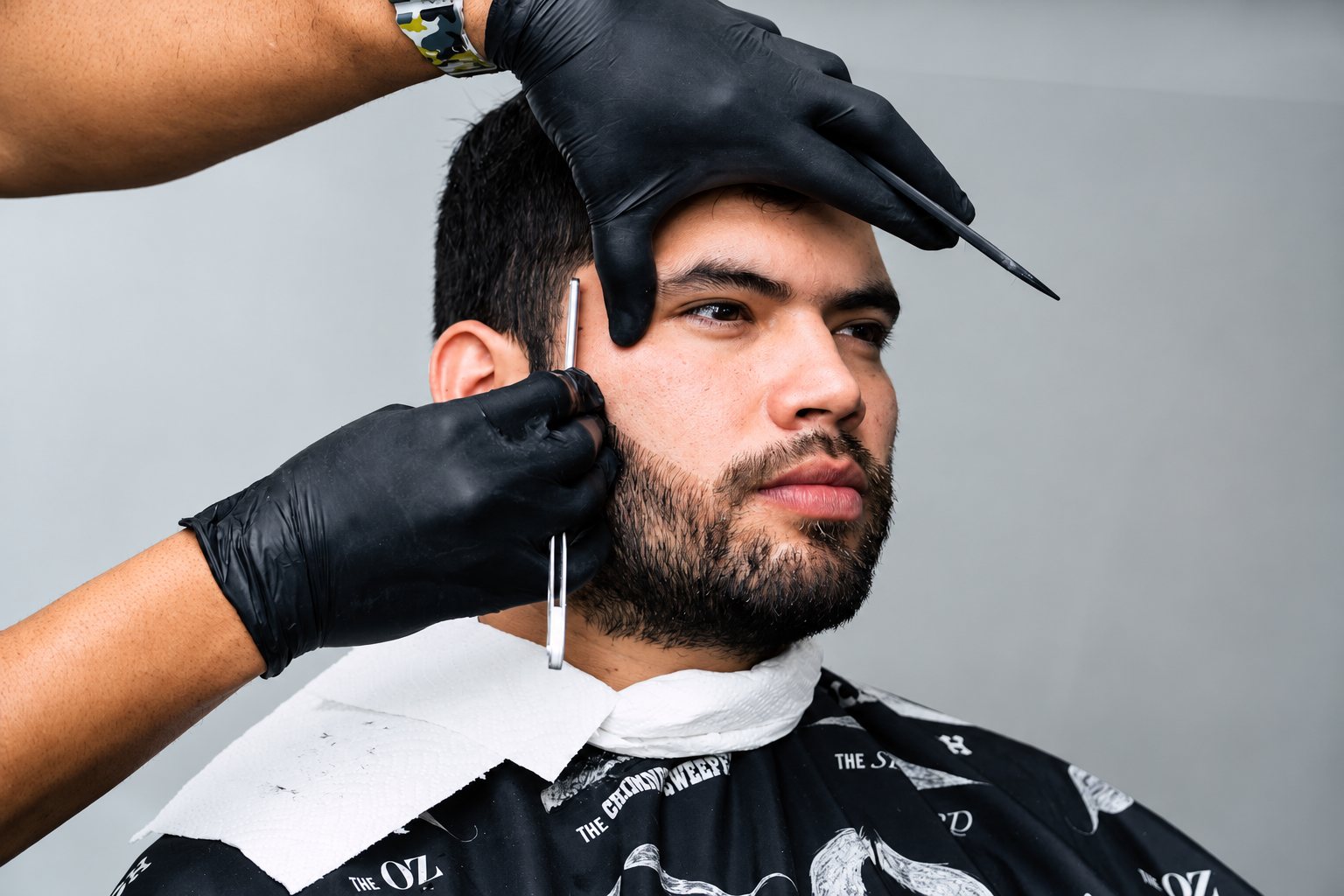 Un barbier ganté de noir taille la barbe d'un client portant une cape noire à motifs, avec des ciseaux et un peigne.