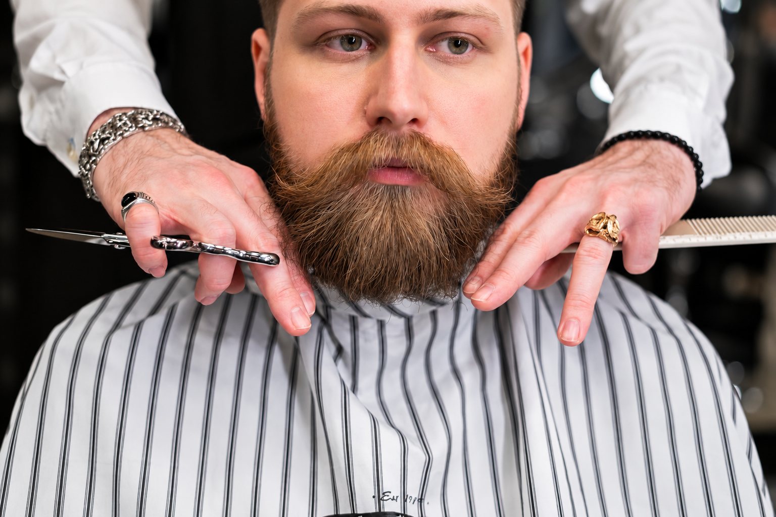 Un barbier tient des ciseaux et un peigne pour tailler la barbe rousse d'un client installé sous une cape rayée.
