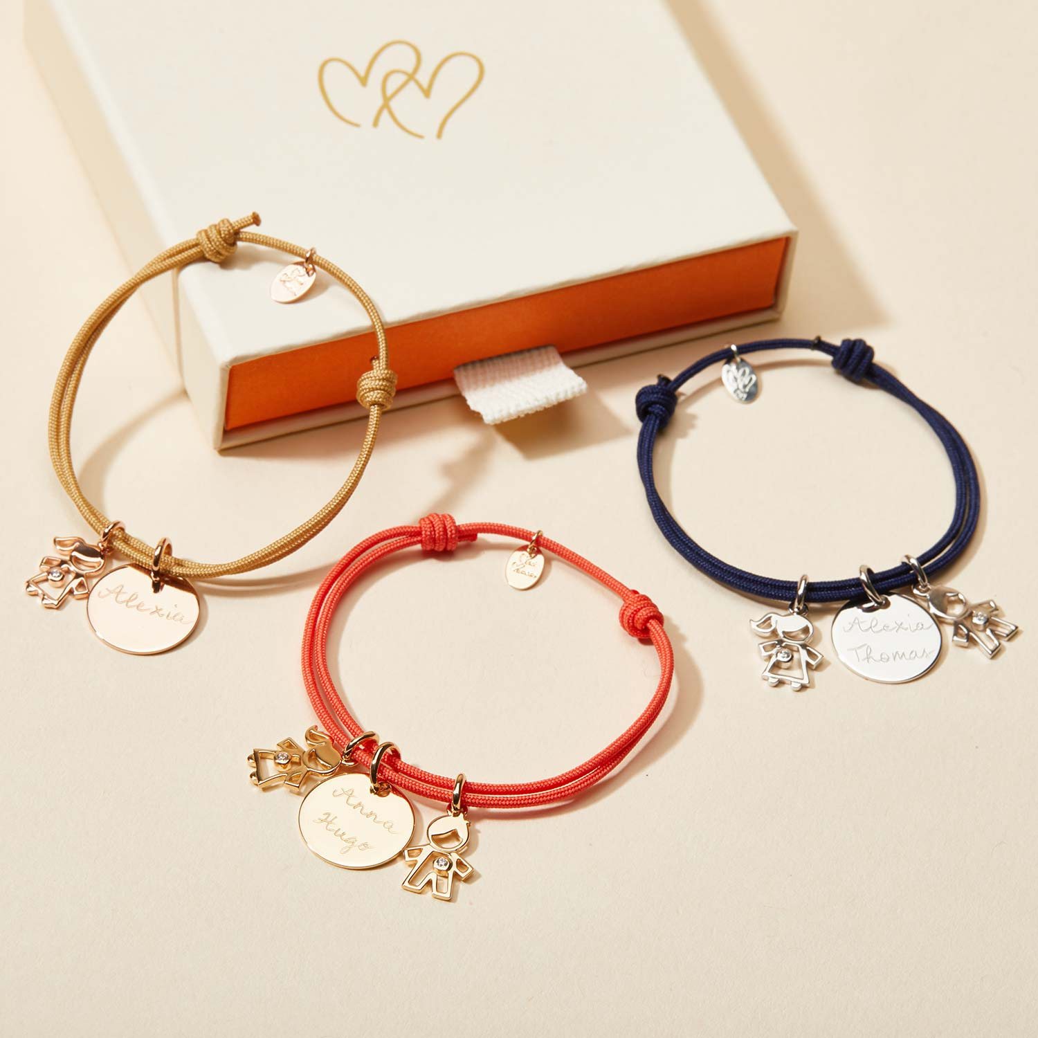 Trois bracelets en cordon beige, corail et bleu marine, ornés de médailles gravées de prénoms et de breloques d'enfants, posés devant un coffret crème.