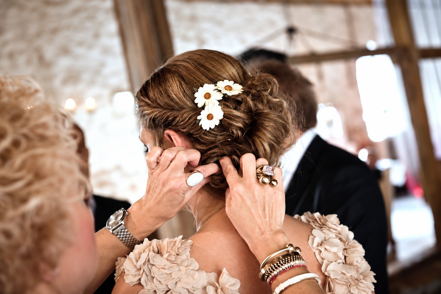 Une femme ajuste de petites marguerites blanches dans le chignon tressé d'une mariée portant une robe à volants en tissu beige.
