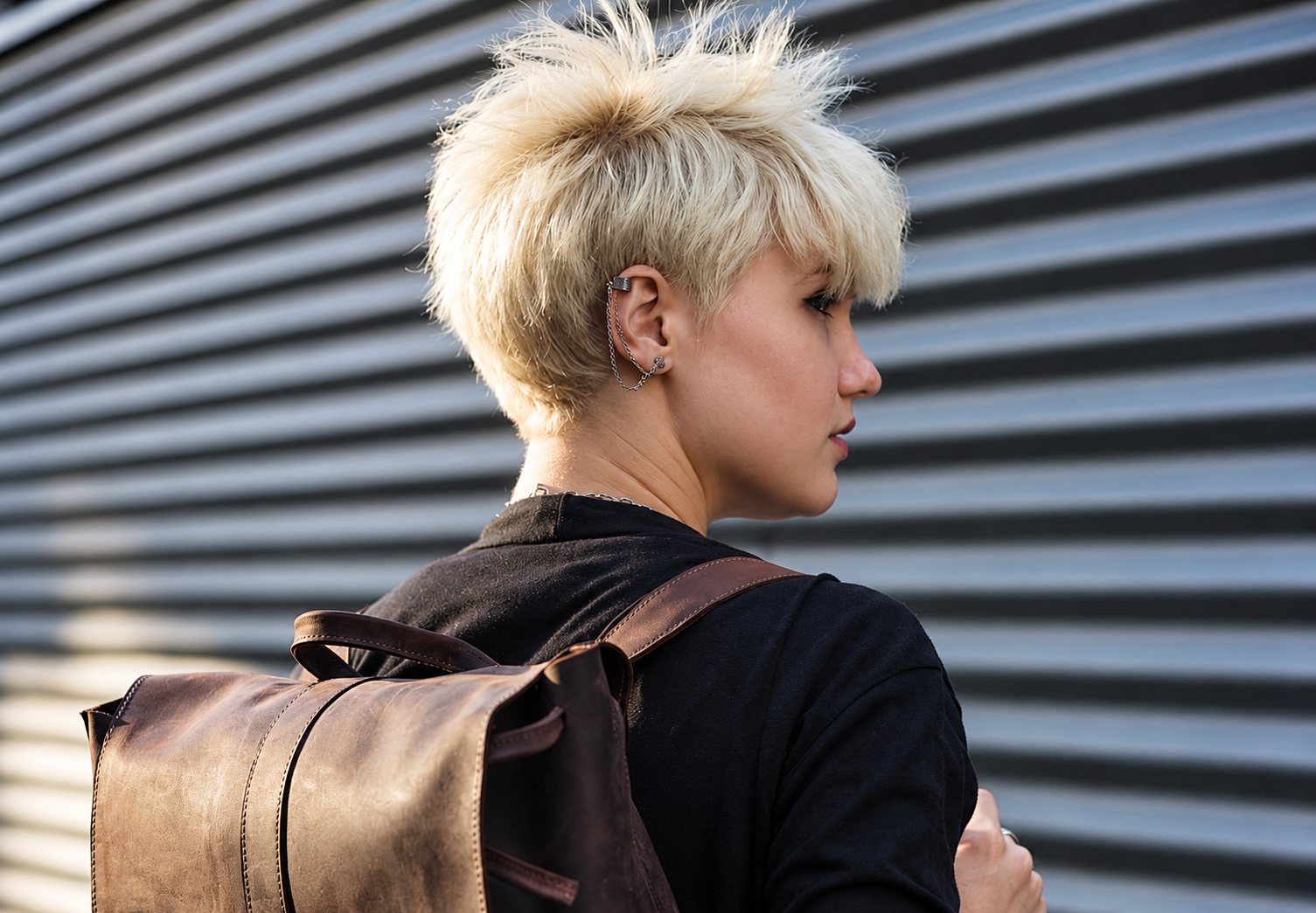 Une femme aux cheveux courts blond platine, vue de profil, porte un sac à dos en cuir marron devant un mur métallique rayé.