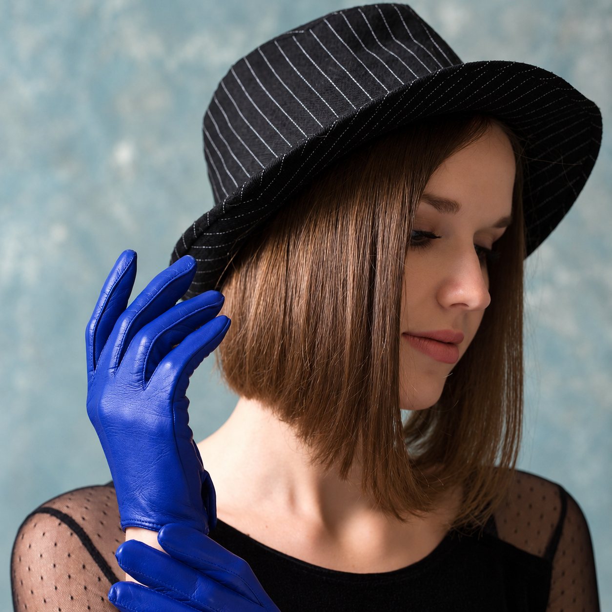 Portrait d'une jeune femme au carré brun, coiffée d'un chapeau noir à fines rayures, qui porte des gants en cuir bleu électrique.