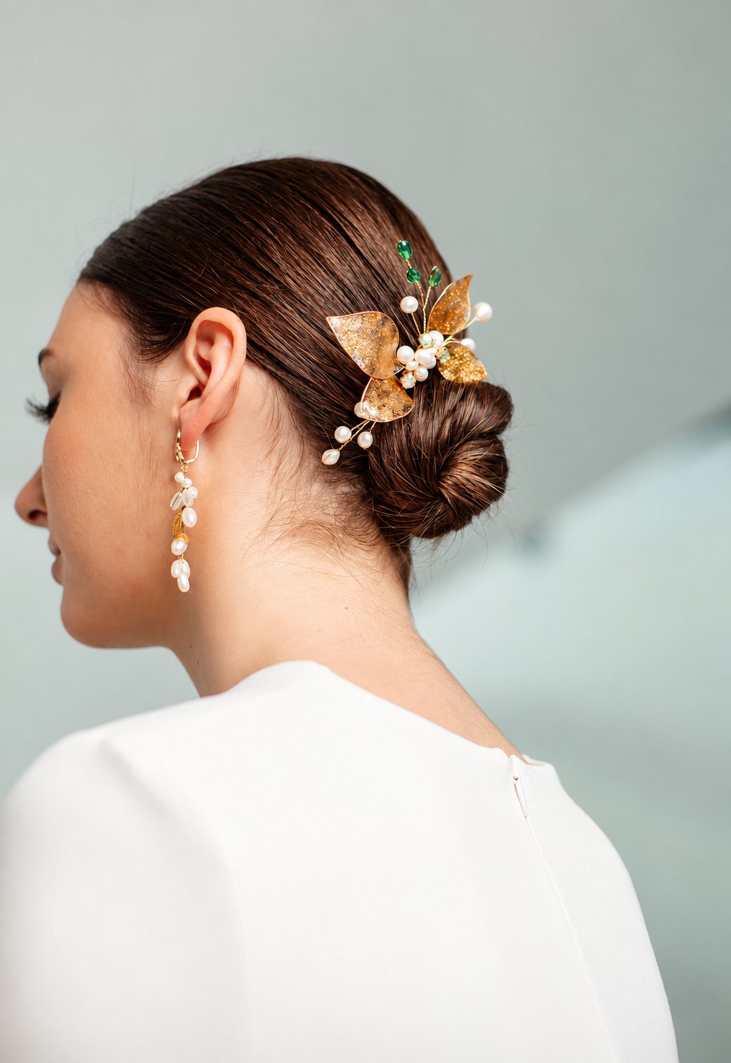 Vue de profil d'une femme au chignon bas orné d'un bijou de cheveux doré à feuilles et perles, avec de longues boucles d'oreilles en perles assorties.