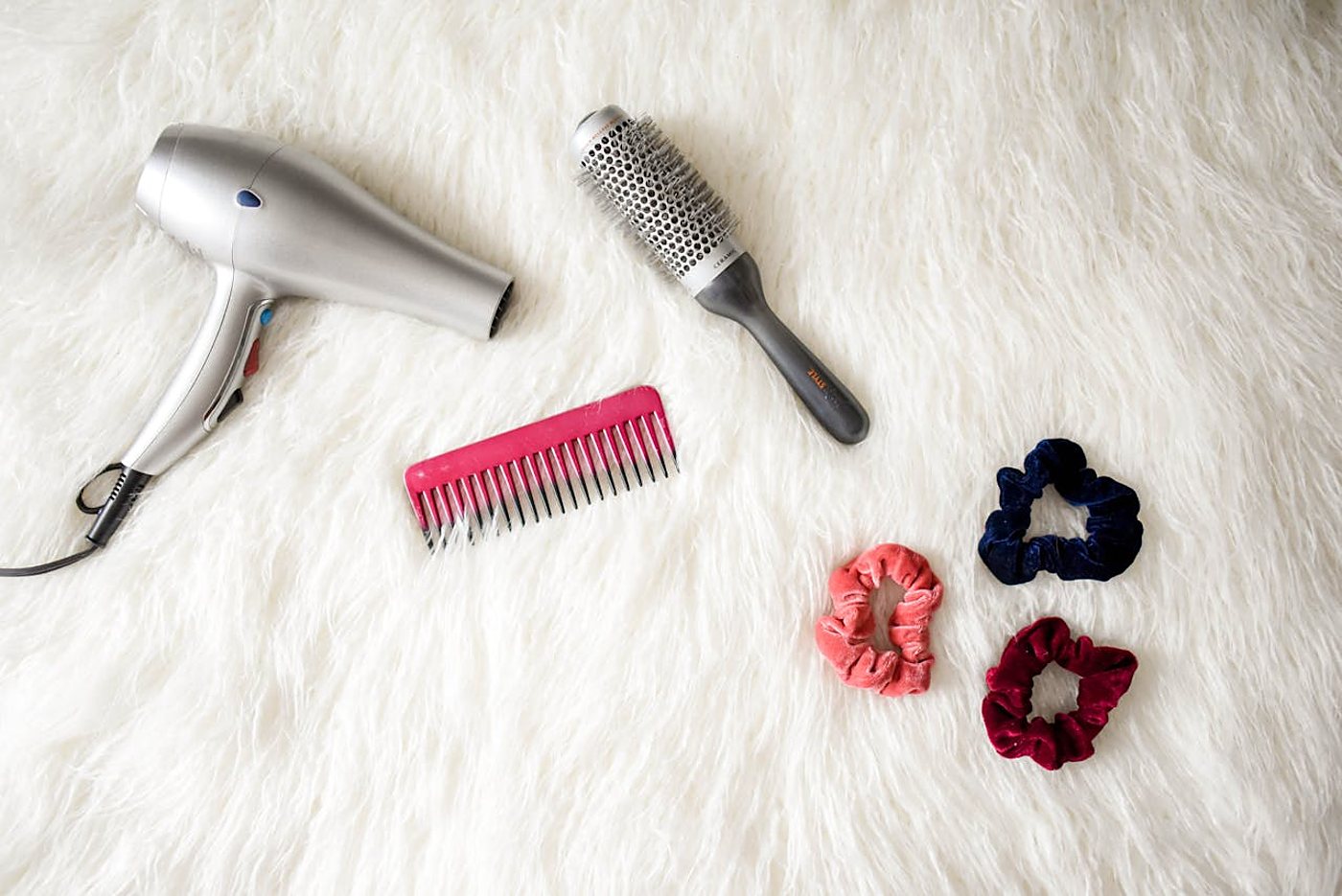Vue de dessus d'un sèche-cheveux argenté, d'une brosse ronde, d'un peigne rose et de trois chouchous en velours posés sur un tapis en fausse fourrure blanche.